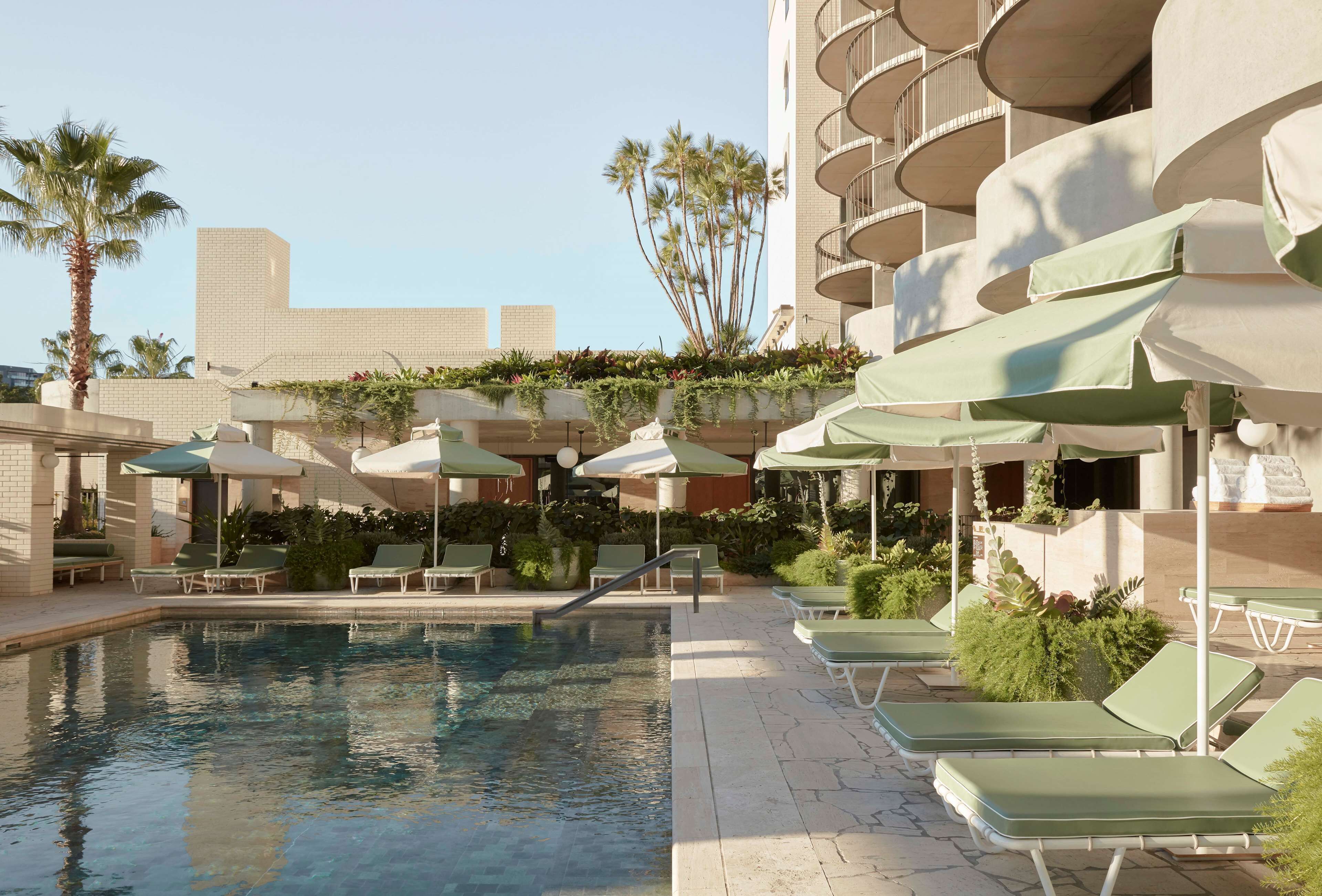 Un espace aménagé au bord de la piscine, avec des chaises longues et des parasols, entouré de verdure et bordé d’un bâtiment doté de balcons.