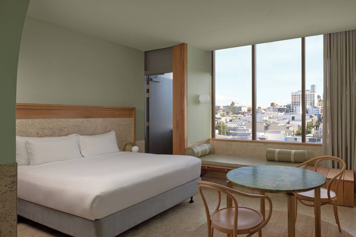 A hotel room with a bed, a seating area with a table, and a large window offering a city view.