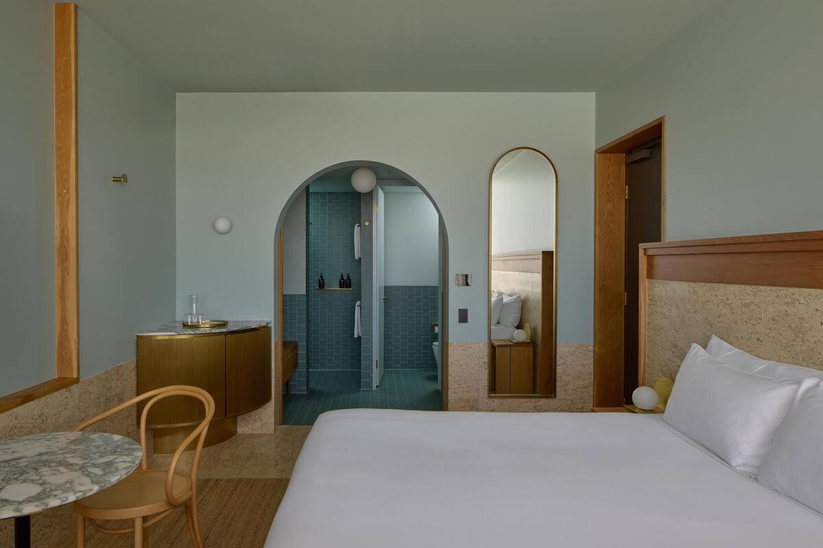 A hotel room with a bed, a round table with a chair, a bathroom with a shower, and a mirror.