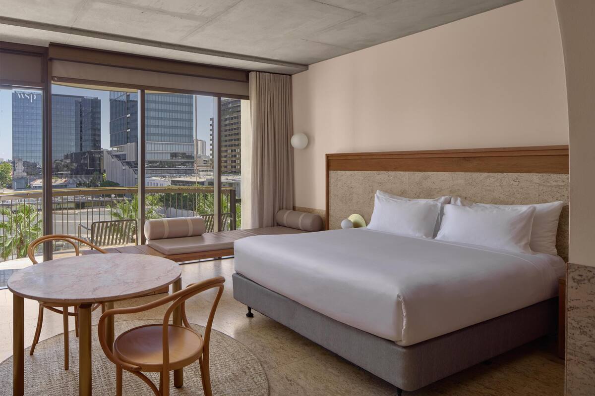 A hotel room with a large bed, a round table, and chairs, a sofa, and a view of city buildings through large windows.