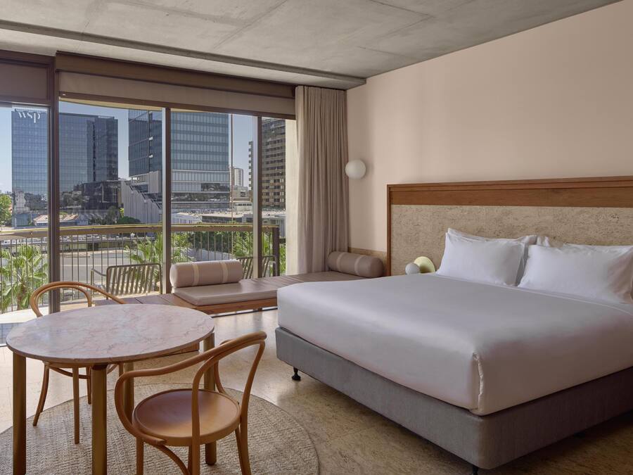Ein Hotelzimmer mit einem großen Bett, einem runden Tisch mit Stühlen, einer Couch und Blick auf Stadtgebäude durch große Fenster.