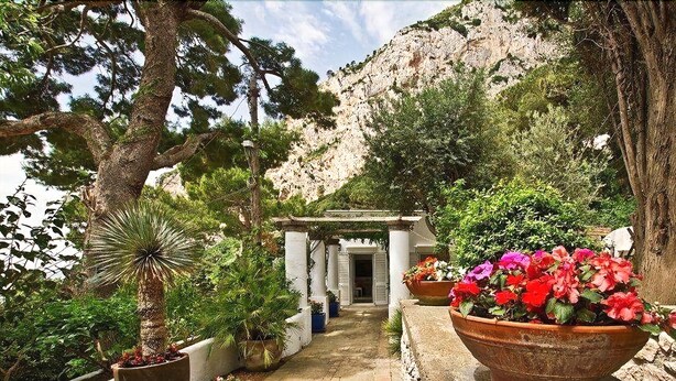 A pathway leading to a building with a white facade and columns, surrounded by potted plants and flowers. - Villa Bougainvillea (Capri)