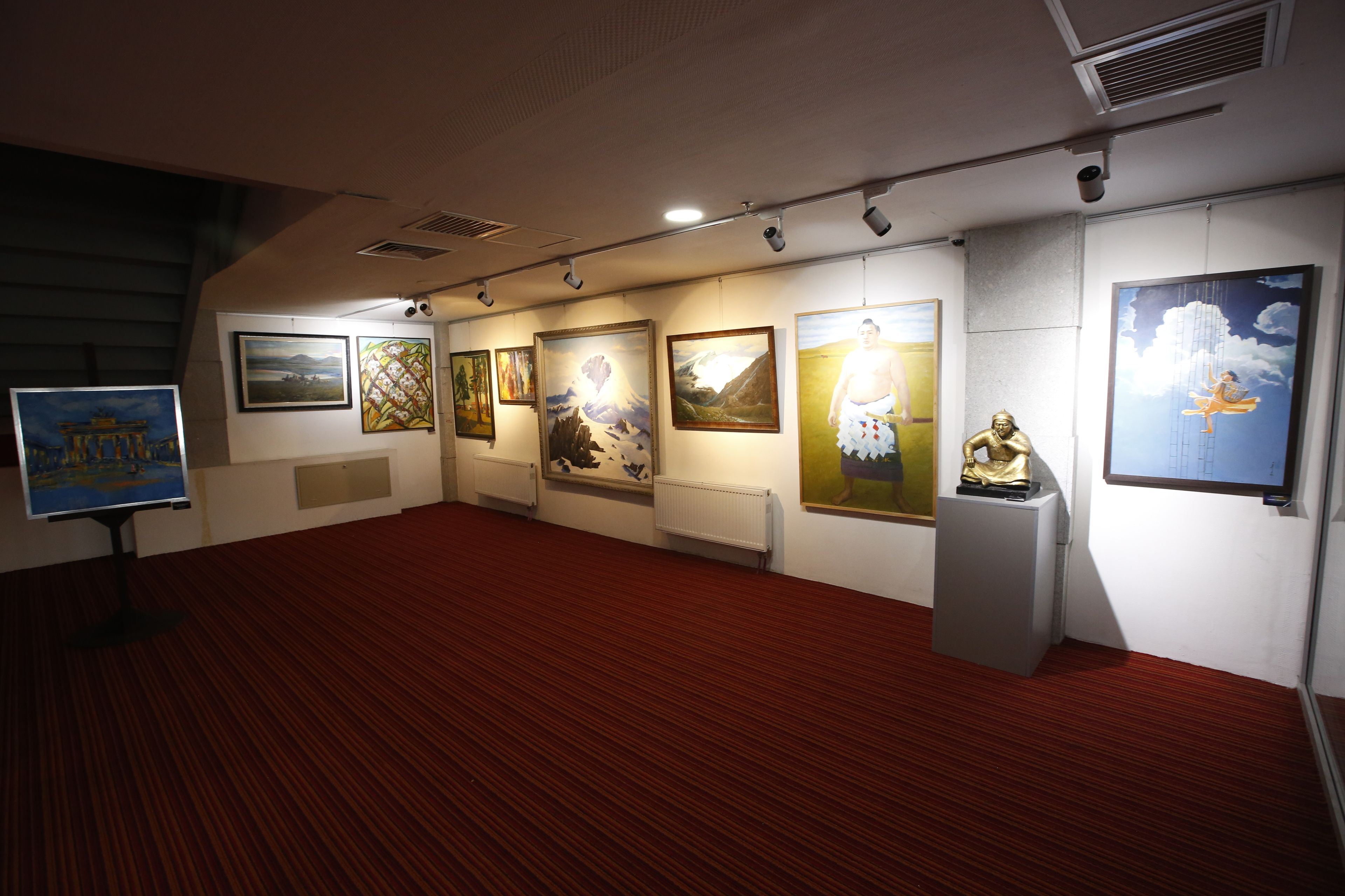 Una galería con obras expuestas, piso cubierto de alfombra roja y pared blanca.
