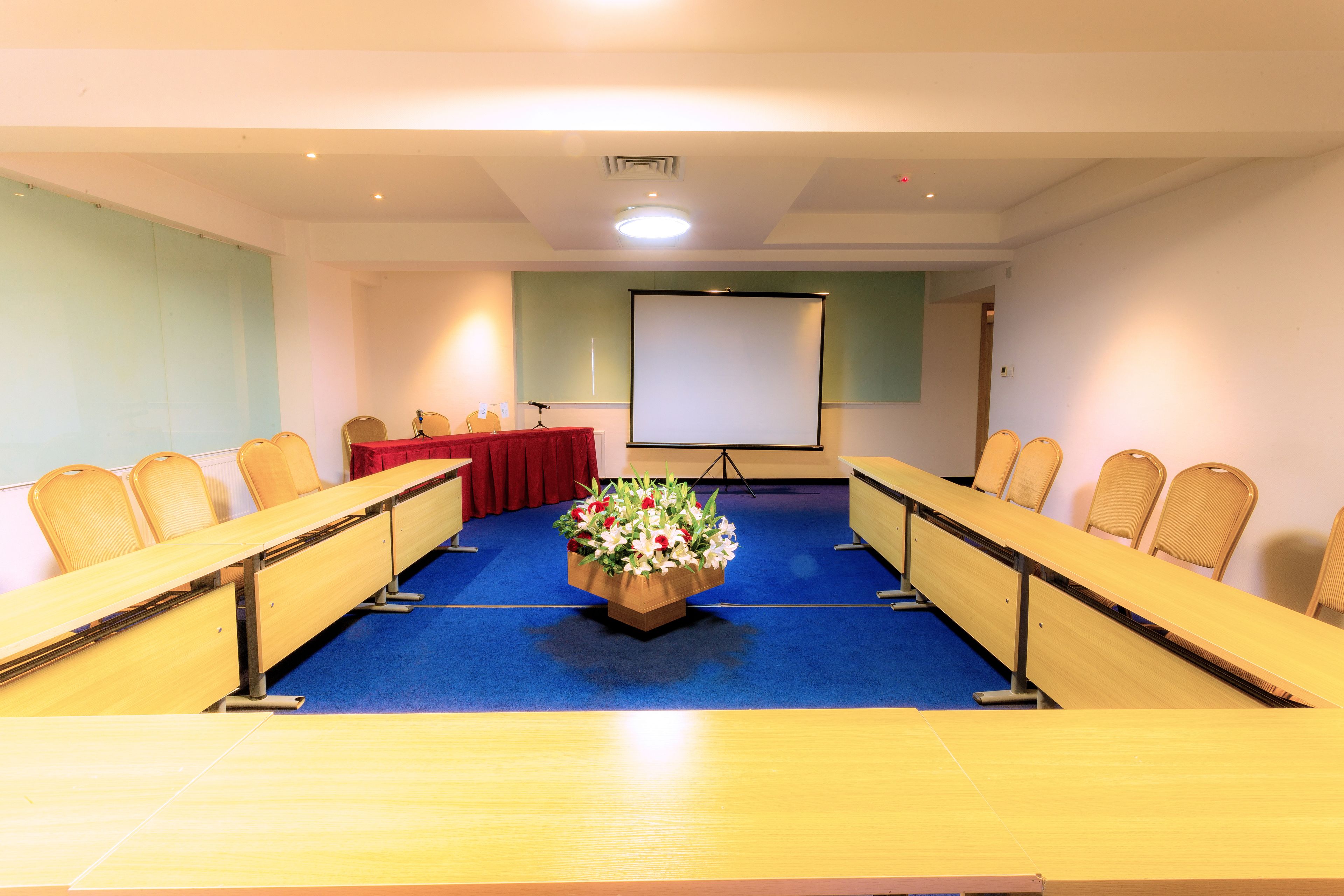 Una sala de conferencias con una larga mesa, sillas, una pantalla de proyector y un arreglo floral.