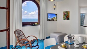 Una habitación con un gran ventanal arqueado con vista al mar, una silla de mimbre, una cama, una mesa con una cafetera y un cuadro.