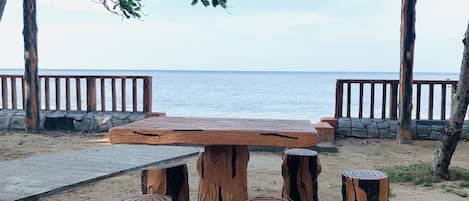Una mesa de madera con cuatro taburetes, ubicada en una playa arenosa con un muro de piedra y baranda de madera, con vista al mar.