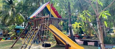 A playground with a slide and climbing structures, surrounded by palm trees and greenery.