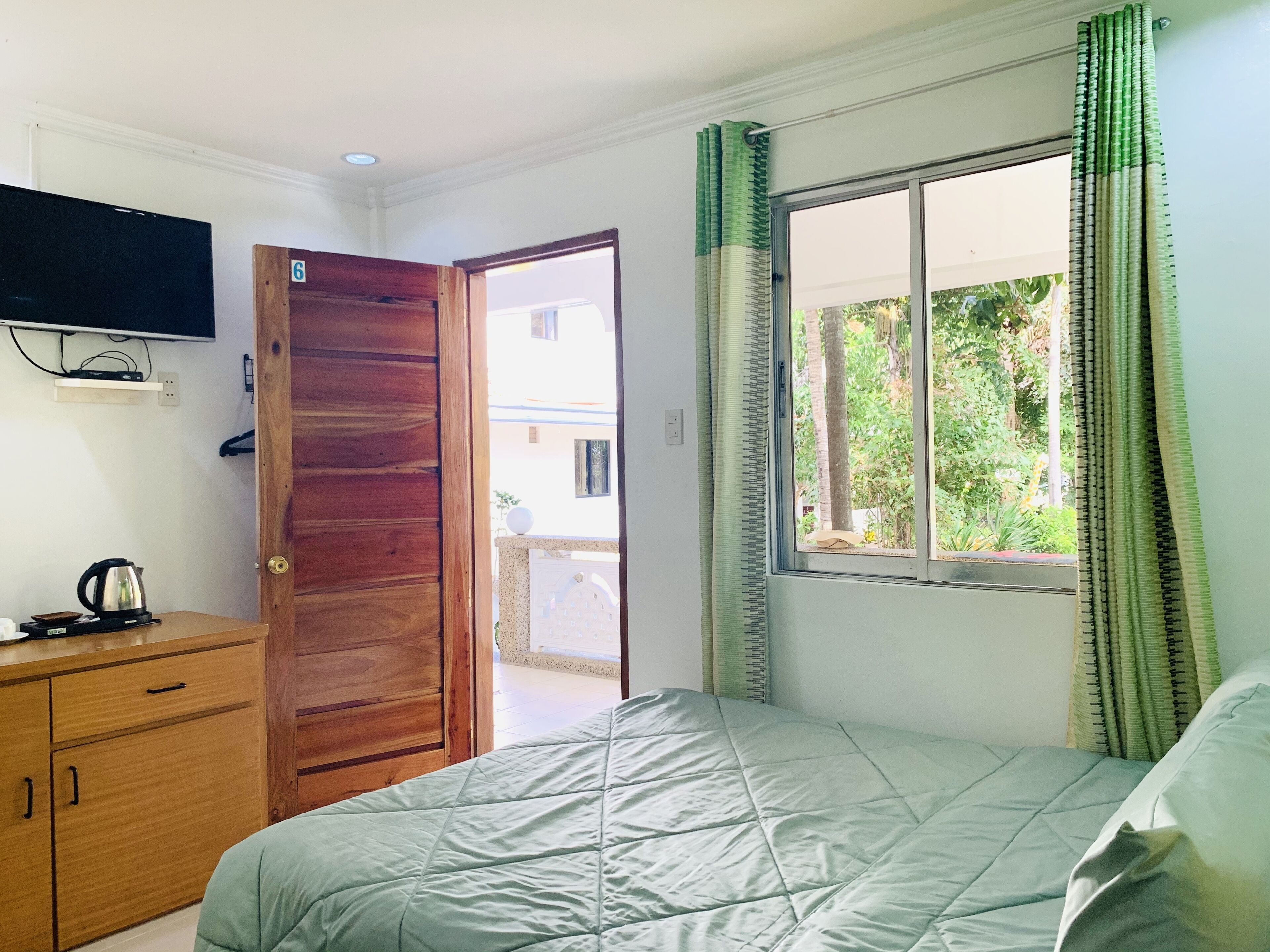 A bedroom with a bed, wooden wardrobe, a television mounted on the wall, and a window with curtains.