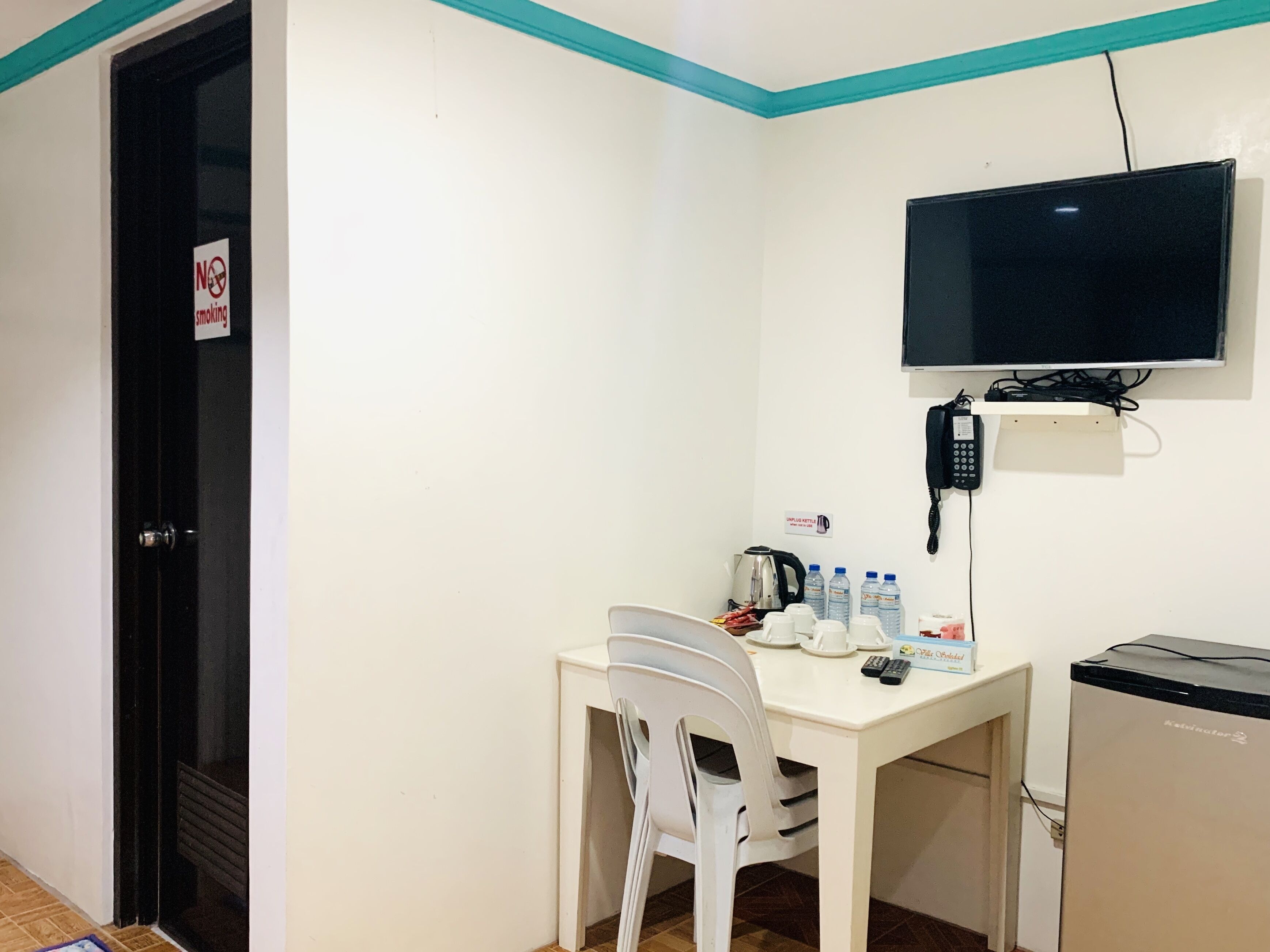 A small, clean room with a white table, a white chair, a television mounted on the wall, a kettle, and a refrigerator.