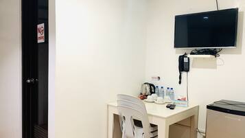 A small, clean room with a white table, a white chair, a television mounted on the wall, a kettle, and a refrigerator.