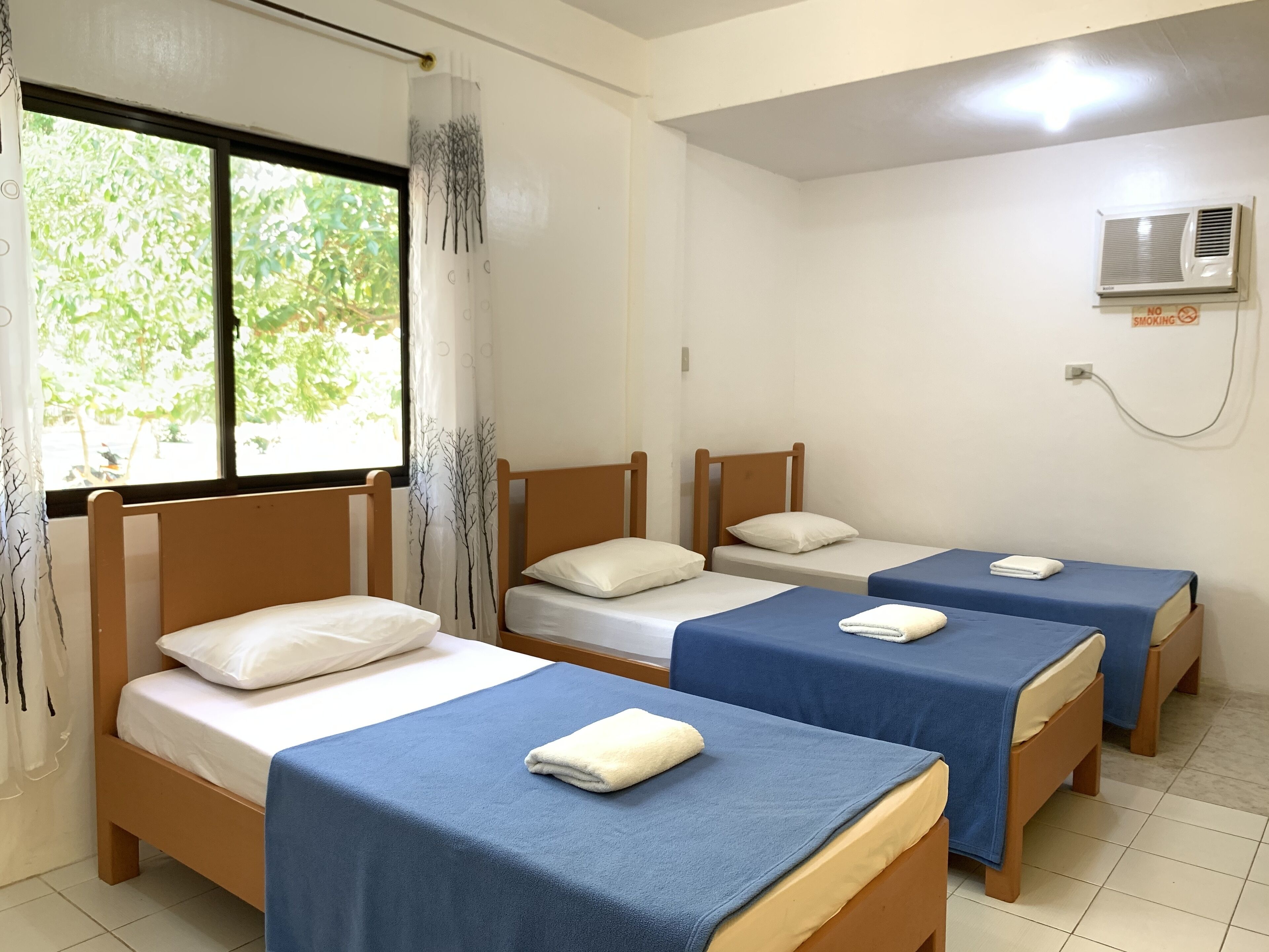 A room with four single beds, each with a blue bedspread and white pillows. There is a window with a view of greenery outside.