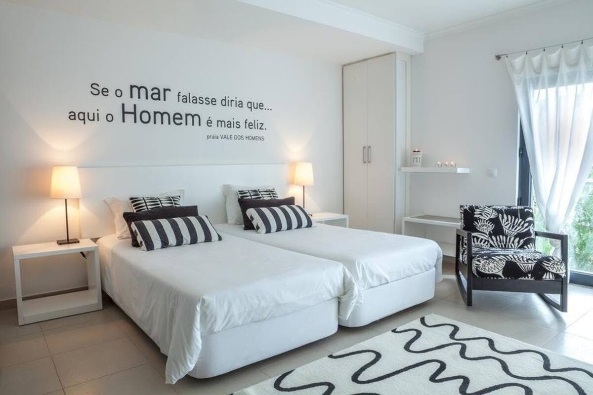 A bedroom with two beds, a wall quote, a chair, and a window with curtains.