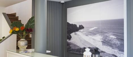 A reception area with a glass counter, a large framed photograph of a beach, a vase with flowers, and a staircase in the background.