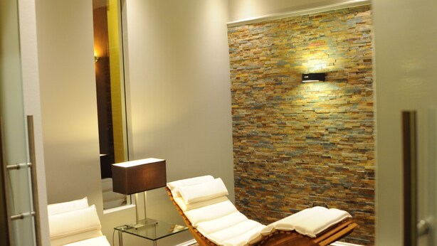 A spa room with two wooden loungers, a glass partition, a stone wall, and a mirror. - Hotel Rheinischer Hof (Erkelenz)