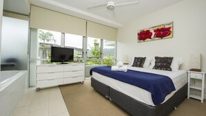 Une chambre moderne équipée d’un lit, d’une télévision, d’un ventilateur de plafond et d’une grande fenêtre donnant sur un paysage verdoyant.