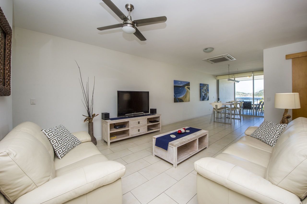 A modern living room with a flat-screen TV, two white leather sofas, a coffee table, and a ceiling fan.