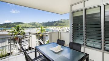 A balcony with a glass table, chairs, and a view of a marina and hills.