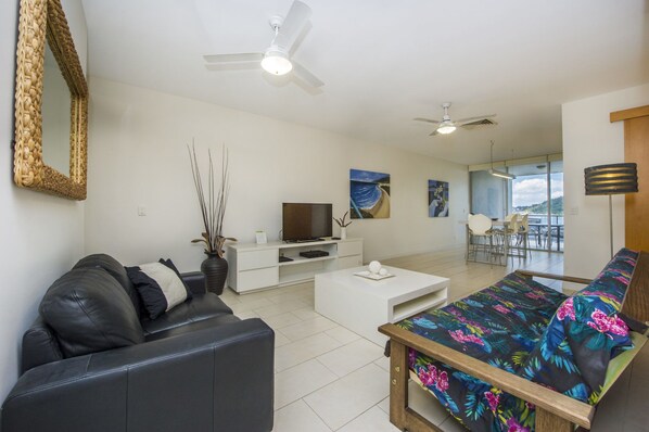 A modern living room with a black leather sofa, a white coffee table, a TV, and a dining area with chairs. - 1 Bright Point Apartment 1405 (Nelly Bay)