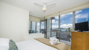 Ein Hotelzimmer mit einem Bett, einer Holzkommode, einem Fernseher und großen Fenstern mit Blick auf das Meer und Gebäude.