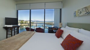 A hotel room with a large bed, a TV, a balcony with a view of the water and boats, and a painting on the wall.