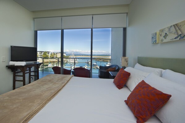 A hotel room with a large bed, a TV, a balcony with a view of the water and boats, and a painting on the wall.