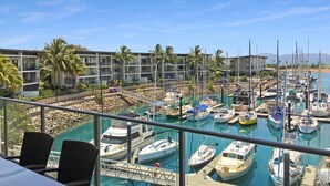 A marina with multiple boats docked, a building with balconies, and a clear blue sky.