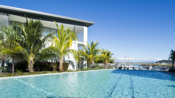 A modern hotel with a swimming pool, palm trees, and a marina in the background.