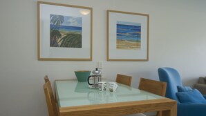 A dining area with a glass-top table, wooden chairs, a blue armchair, and two framed beach paintings on the wall. - Blue on Blue Superior Studio Room 1241 (Nelly Bay)