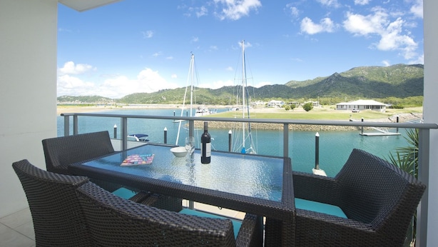 A balcony with wicker furniture, a glass table, and a view of a marina with boats and hills in the background. - Blue on Blue Superior Studio Room 1241 (Nelly Bay)