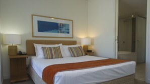A neatly made bed with white linens and a brown throw blanket, two striped pillows, and a framed picture above it. There are two bedside tables with lamps, a wicker basket, and a telephone. - Blue on Blue Superior Studio Room 1241 (Nelly Bay)