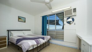 Quarto com cama, janela com cortinas, ventilador de teto e ar condicionado, além de um pequeno quadro na parede.