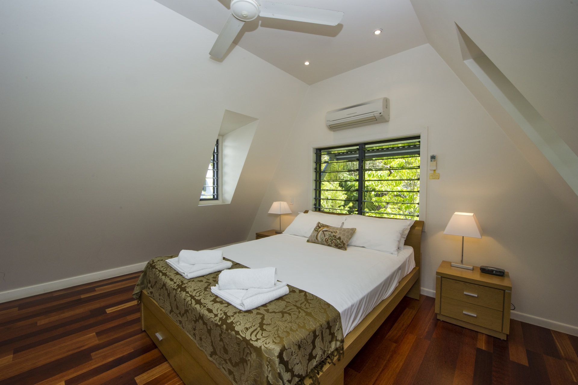 A bedroom with a bed, a nightstand, a ceiling fan, an air conditioning unit, a window with blinds, and a small skylight.