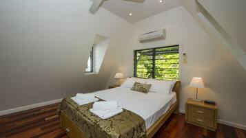 A bedroom with a bed, a nightstand, a ceiling fan, an air conditioning unit, a window with blinds, and a small skylight.