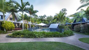 A pool surrounded by lush greenery and palm trees, with a covered seating area and a building in the background.