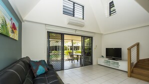 Una sala de estar con un sofá de cuero negro, un televisor sobre un mueble blanco y un ventanal corredero que da a un patio exterior.