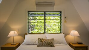 Un dormitorio con cama, dos mesitas de noche, una ventana con vistas a zonas verdes y un equipo de aire acondicionado sobre la ventana.