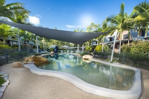 A swimming pool with a curved edge, surrounded by palm trees and a shaded area. - Canopy Chalet 10 (Nelly Bay)