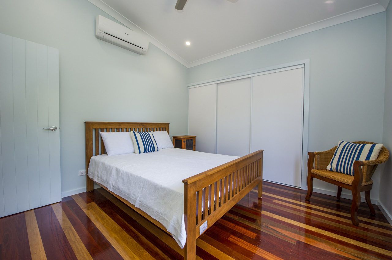 Quarto com cama de madeira, paredes brancas, porta branca, guarda-roupa branco e cadeira de vime.