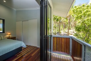 Une chambre à coucher comprenant un lit, une table de chevet, une lampe et une porte coulissante donnant sur un balcon avec vue sur les arbres.