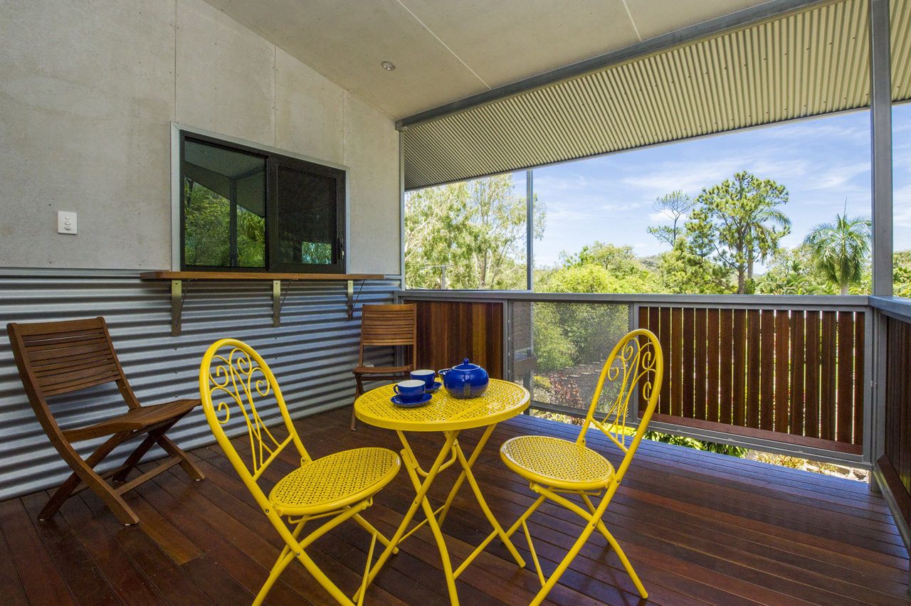 Área externa coberta com deck de madeira, cadeiras amarelas e uma mesa com copos azuis.