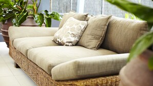 Un sofá de mimbre con cojines color beige y un cojín estampado, ubicado en una habitación con ventanas amplias y plantas en macetas.