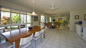 A spacious dining area with a wooden table and white chairs, large windows, and a ceiling fan. - John's Tropical Island Home (Nelly Bay)