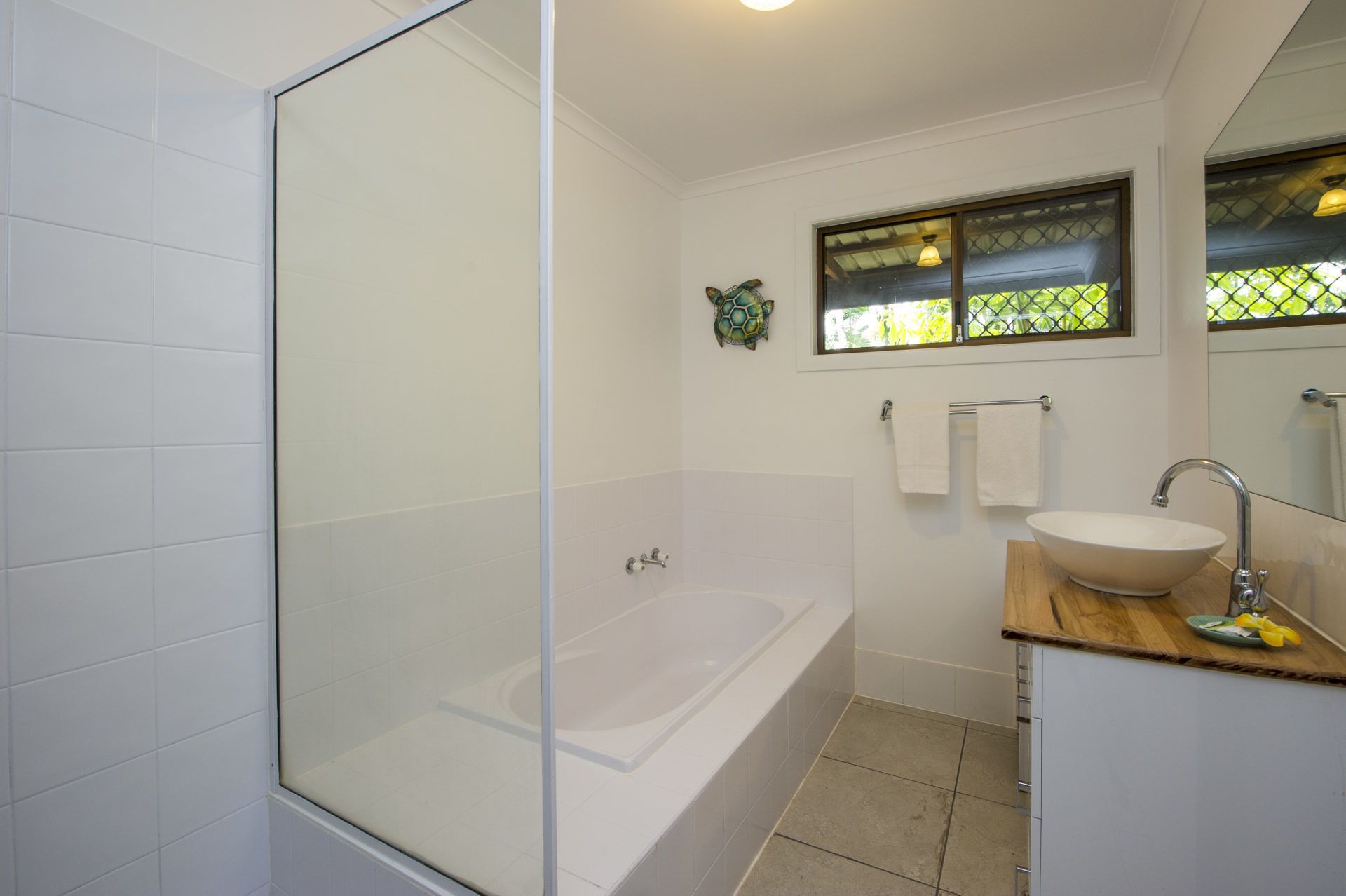A modern bathroom with a glass-enclosed shower, a freestanding bathtub, a wooden vanity with a vessel sink, and a window with a view of greenery.