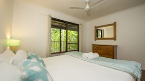 Un dormitorio con cama, mesita de noche, lámpara, ventilador de techo, ventana con vistas al verdor y cómoda de madera.