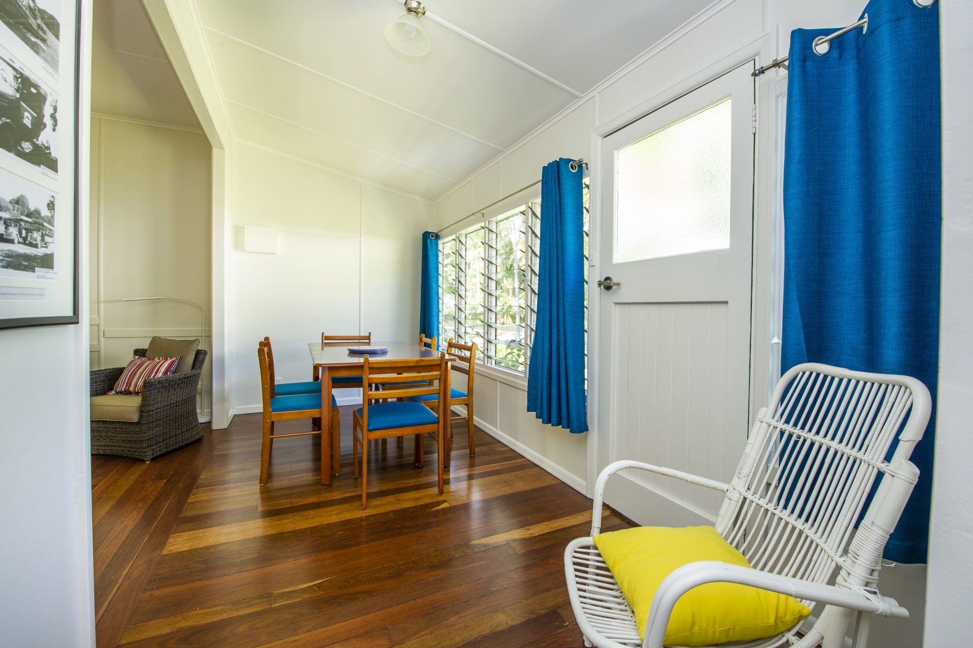 A room with a wooden floor, a dining table with chairs, a white chair with a yellow cushion, and blue curtains.