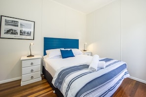 Una cama bien hecha con un cabecero azul, ropa de cama blanca y cubrecama azul y blanco en el pie de la cama. Hay dos mesitas de noche con lámparas, un aparador blanco y un cuadro en la pared.