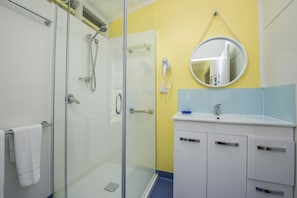 Un baño con cabina de ducha de vidrio, un lavabo blanco con lavamanos azul y un espejo redondo en la pared.