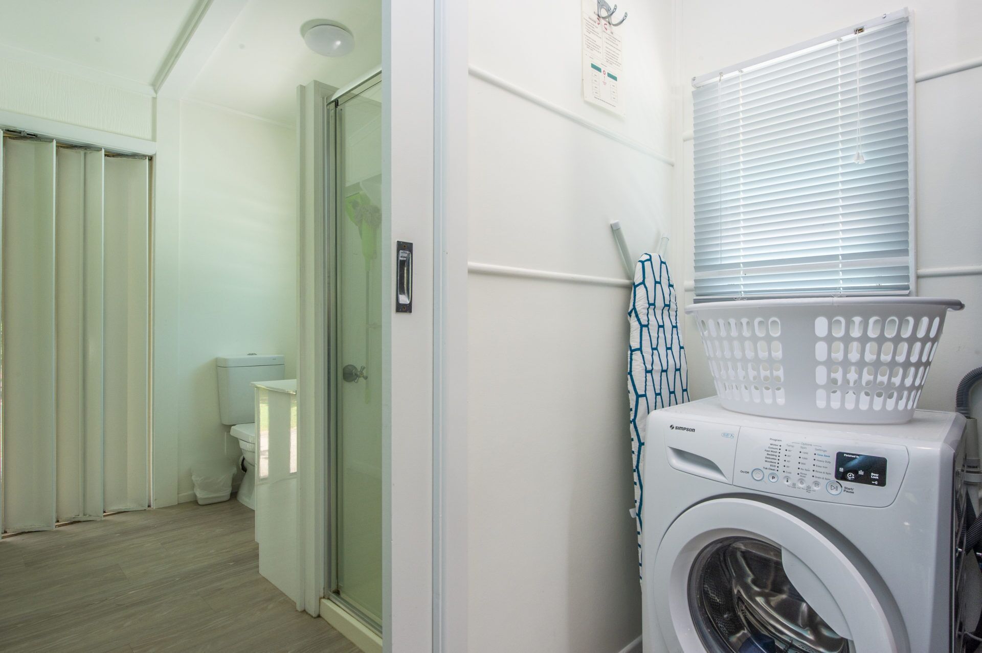 A laundry room with a washing machine, a glass shower enclosure, and a window with blinds.