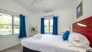 Una habitación de hotel con cama, mesita de noche, lámpara, estantería y ventana con cortinas.