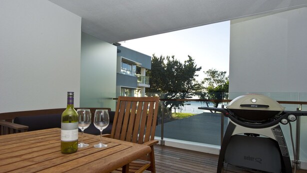 Una mesa de madera con una botella de vino y copas, una parrilla y vistas a un edificio y árboles.
